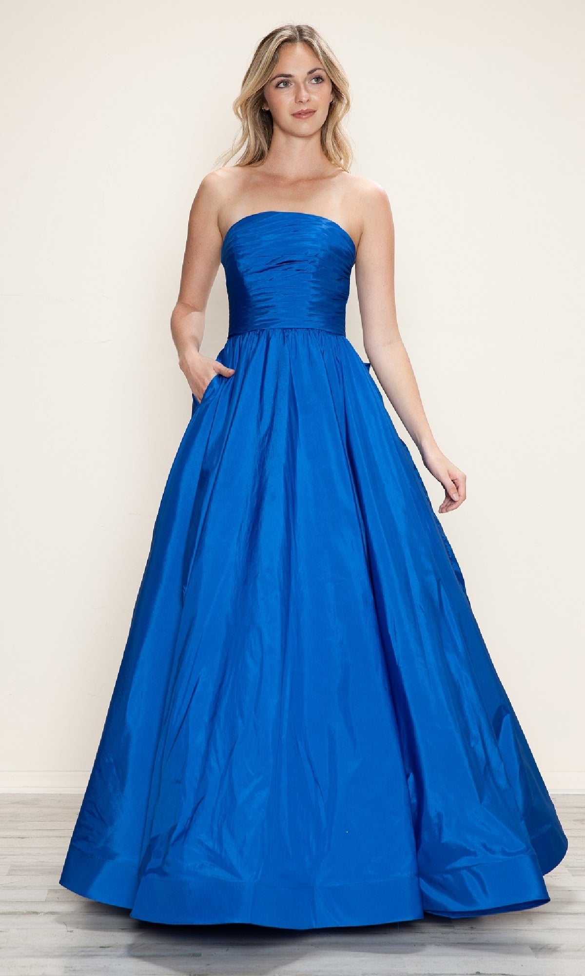 A woman in the Long Prom Dress: Poly USA 9754, featuring a strapless ruched bodice and floor-length bright blue taffeta skirt with an adjustable lace-up back, stands on a wooden floor against a plain light background.